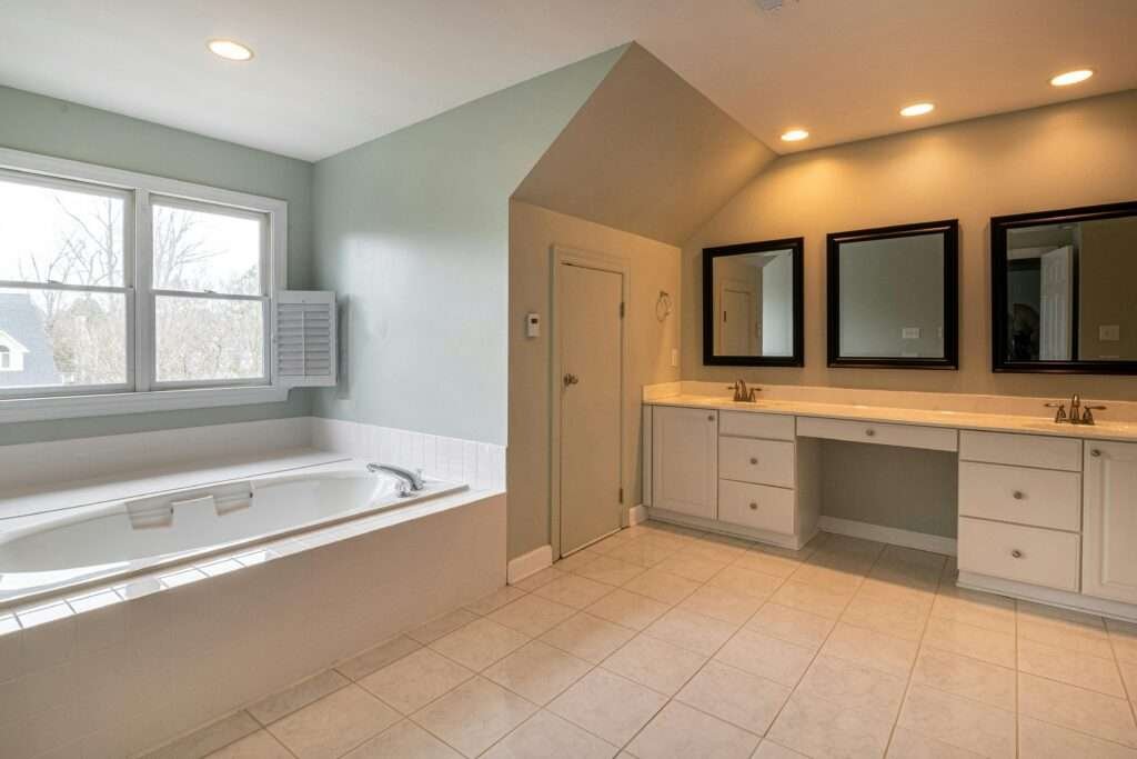 A large bathroom featuring a built-in bathtub, a long double vanity with two sinks and mirrors, and tiled flooring. The room has light green walls and recessed lighting, creating a bright and airy atmosphere. Location: Wolverhampton, United Kingdom